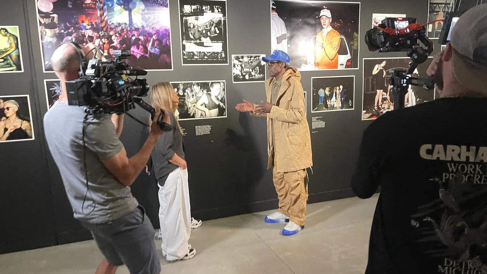 A camera operator and a second shooter film an interview in front of a wall of 1990s club photographs
