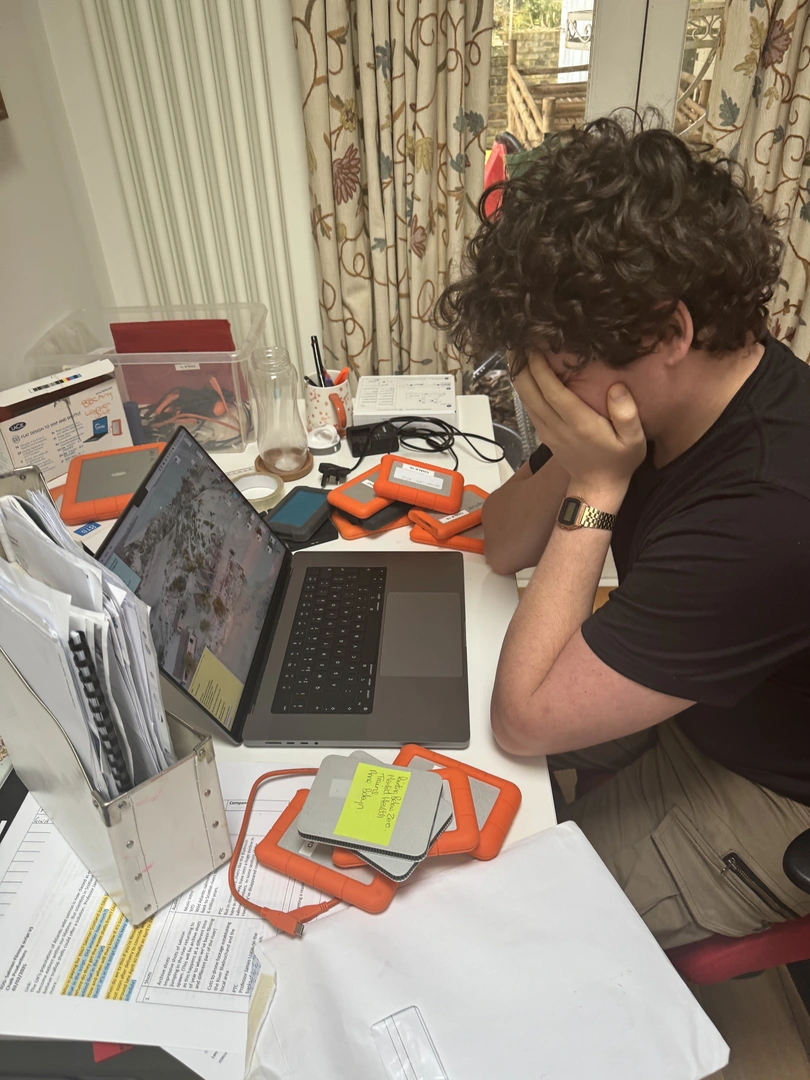 A researcher surrounded by hard drives, papers, and a laptop — the reality of managing production media across too many tools